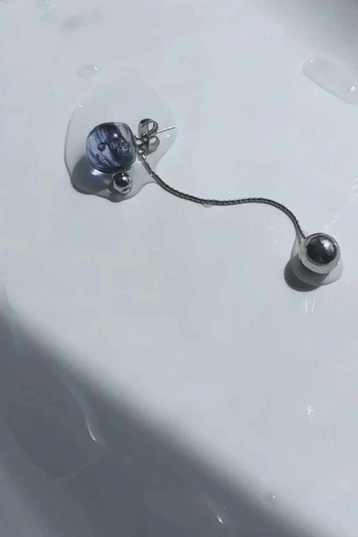 A video of the SORA short drop earring with detachable chain—features a hand-molded blue orb that shifts in tone like the sky, shown laid on neutral surface.